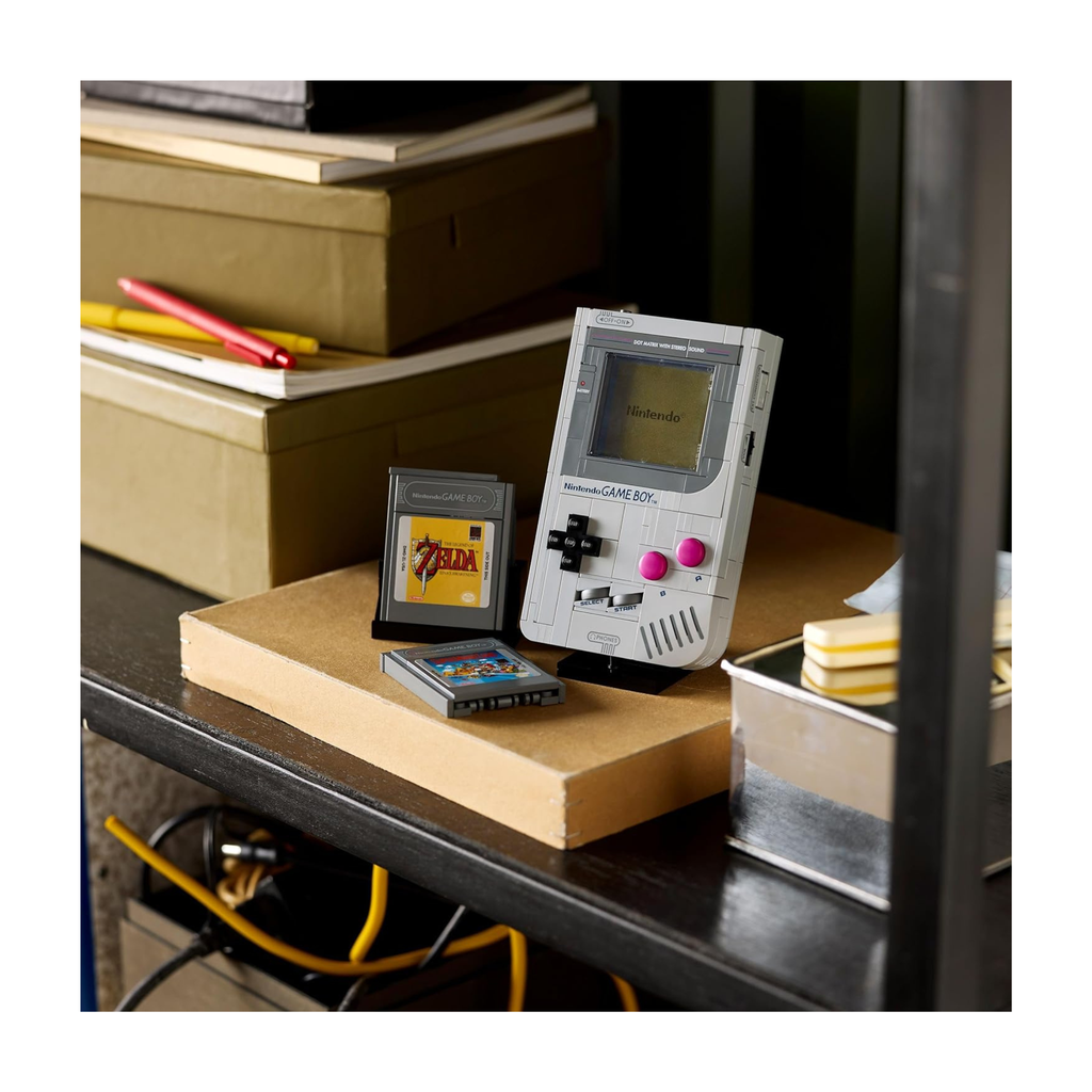 Handheld gaming device resembling a Game Boy on a shelf with two game cartridges and stationery items in the background.