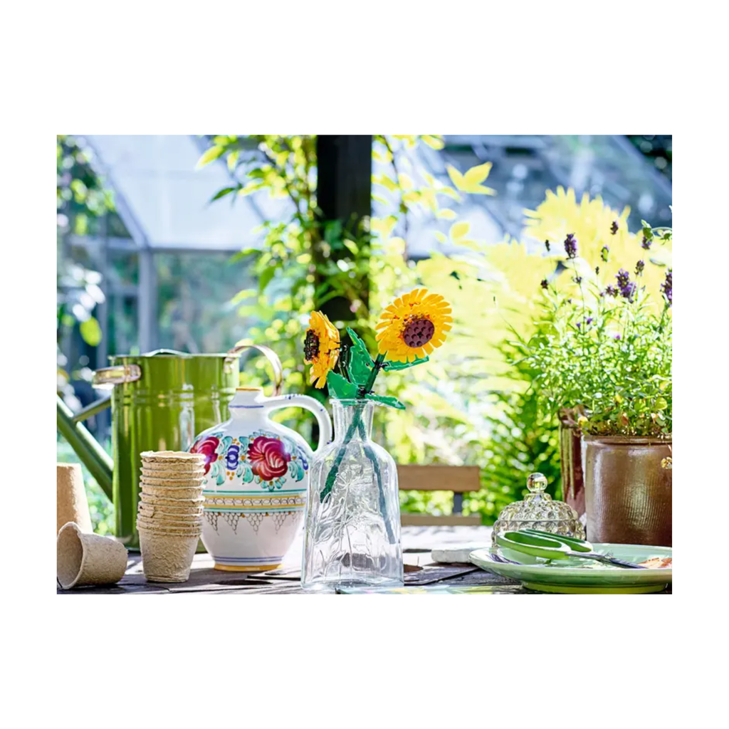 Two vibrant LEGO Sunflowers in a clear vase with green stems, surrounded by decorative items and greenery.