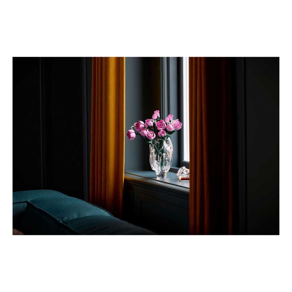 Bouquet of pink roses in a clear vase with green foliage, set against a dark wall and window curtains.