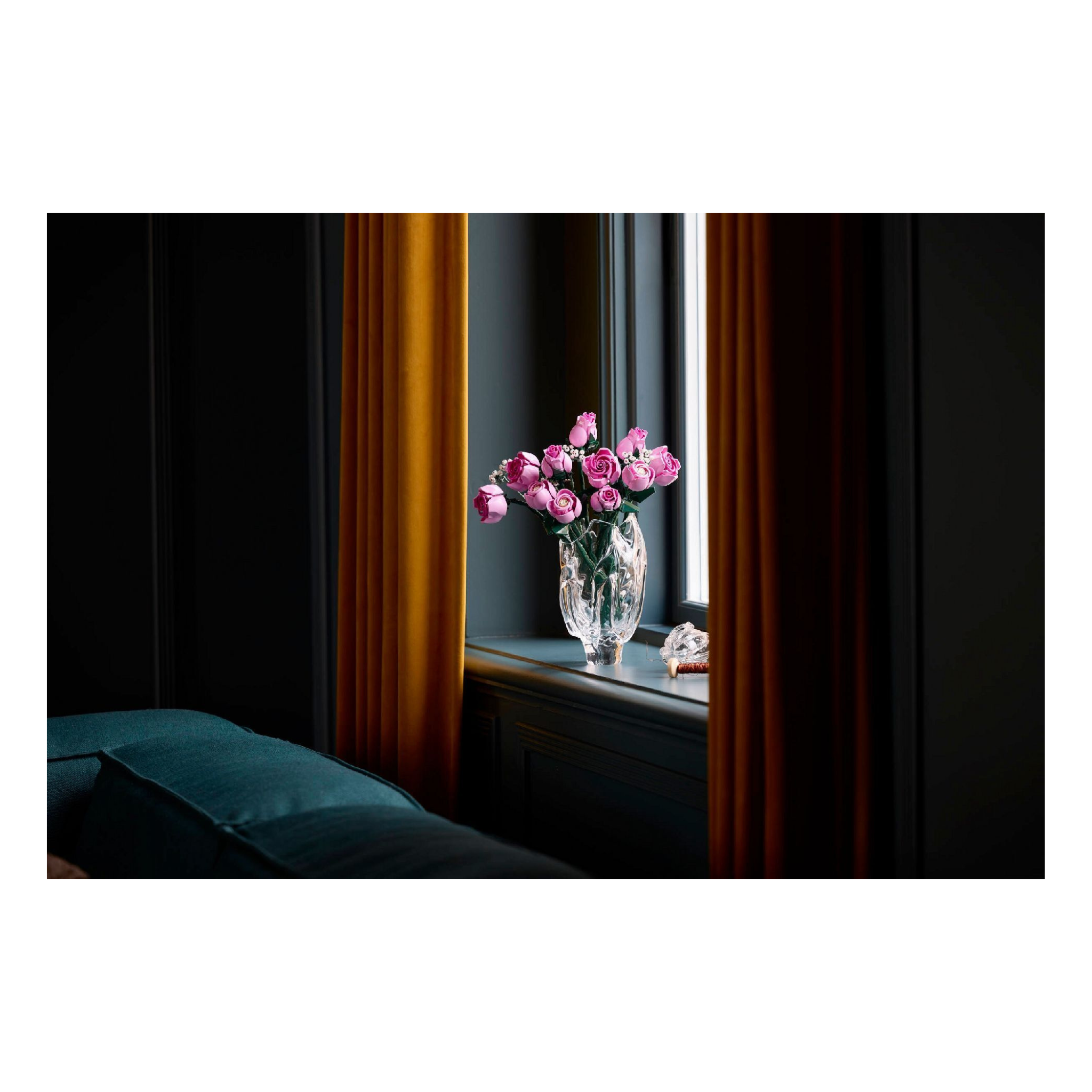 Bouquet of pink roses in a clear vase with green foliage, set against a dark wall and window curtains.