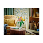 Colorful bouquet featuring pink daisies and yellow blooms in a clear vase, set on a wooden table with art supplies in the background.