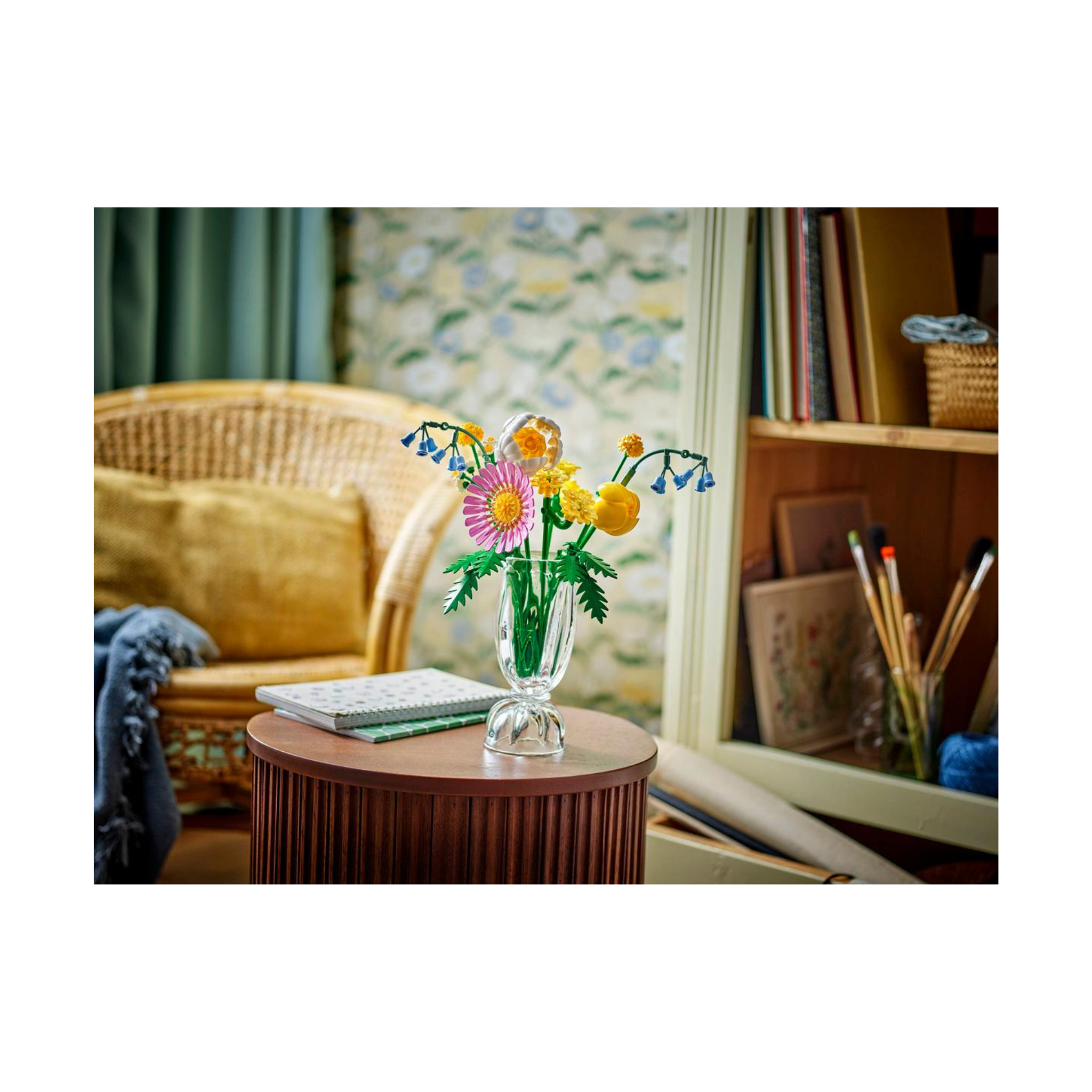 Colorful bouquet featuring pink daisies and yellow blooms in a clear vase, set on a wooden table with art supplies in the background.